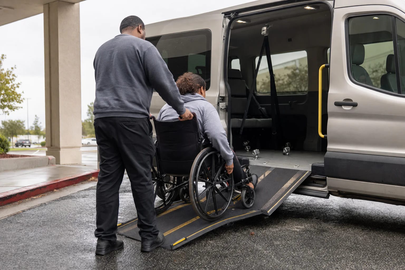 Driver secures a passenger wheelchair inside an unbranded accessible medical transport van at an outpatient clinic curb